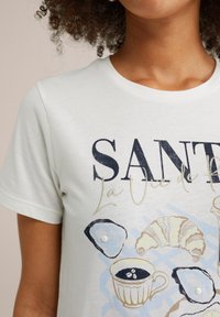 Light grey t-shirt featuring a graphic print of a coffee cup, oysters, and a croissant in blue and neutral hues with pearl accents.