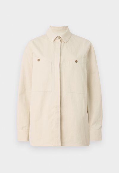 Beige button-up shirt made of cotton with a pointed collar, two front pockets, and brown button accents. Long sleeves with buttoned cuffs.