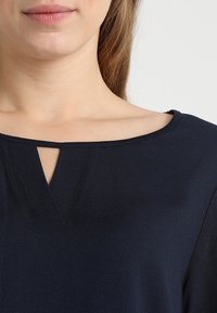 Femme portant un haut bleu marine avec une encolure en V et des manches longues, dévoilant une partie du cou et des lèvres.