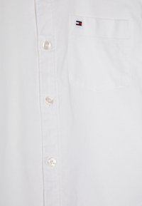 Camicetta bianca con bottoni, caratterizzata da una sola tasca sul petto, bottoni in plastica e una piccola etichetta con logo in rosso, bianco e blu sulla tasca.