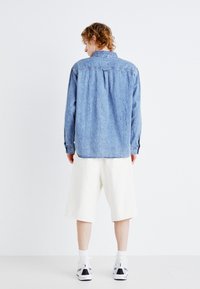 Cămașă din denim de culoare albastru deschis, cu yoke la spate, manșete cu nasturi și plinsuri laterale. Asortată cu pantaloni scurți albi și pantofi sport negri.