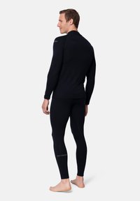 Uomo che indossa una maglia termica nera a maniche lunghe e leggings, a piedi nudi, in piedi leggermente di lato su uno sfondo bianco.