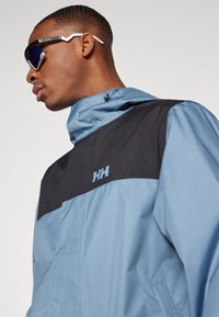 Light blue jacket with black shoulder panels, featuring a zip front, hood, and embroidered logo. Accessories include reflective sunglasses.