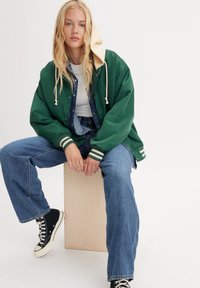 Zielona kurtka bomberka z beżowym kapturem, białymi sznurkami i paskowanymi mankietami, w połączeniu z niebieskimi dżinsami i czarnymi sneakerami za kostkę.