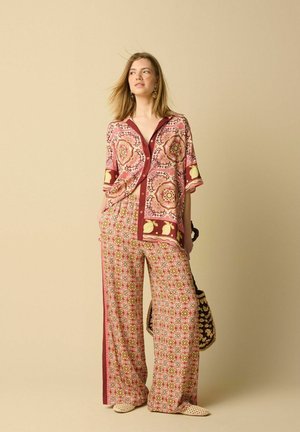 Vrouw draagt een gebloemd overhemd met knopen en bijpassende wijde broek, houdt een geweven tas vast en staat tegen een beige achtergrond.