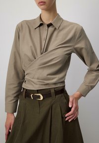 Khaki wrap shirt dengan kerah dan penutupan kancing, dipadukan dengan rok hijau zaitun dan ikat pinggang cokelat dengan gesper logam emas.