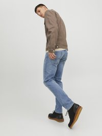 Homme portant une veste marron, un jean bleu clair et des chaussures noires, se retournant avec un pied levé devant un fond clair.