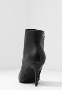 Schwarze Leder-Stiefelette mit hohem Absatz und schlankem Stilettostiefel, mit einem Reißverschluss hinten, von hinten auf einer weißen Oberfläche gezeigt.