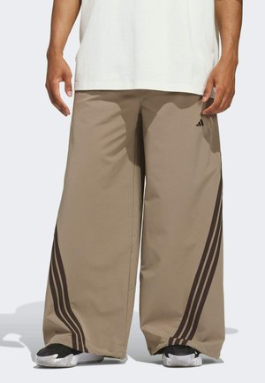 Homme portant un pantalon beige à jambes larges avec des triples rayures foncées sur les côtés et des baskets noires, debout les mains détendues le long du corps.