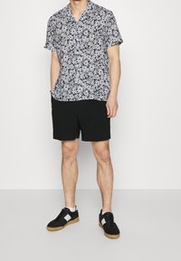 Hombre vestido con una camisa de manga corta negra y blanca con estampado floral, pantalones cortos negros y zapatillas negras con calcetines blancos, de pie contra un fondo blanco.