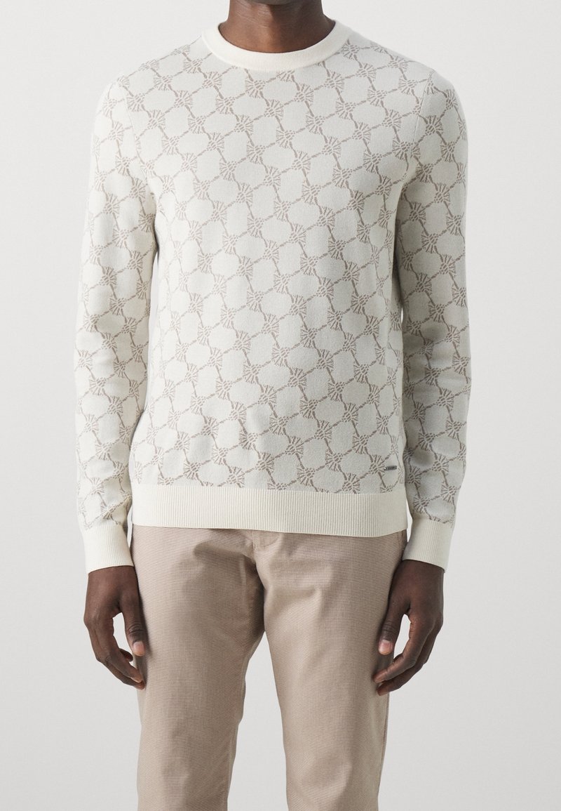 Strickpullover in Creme mit einem strukturierten Muster aus ineinander verflochtenen Linien. Gerippte Bündchen und Saum, kombiniert mit hellbeigen Hosen.