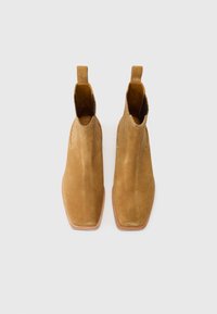 Botas de tobillo de ante en tono beige con punta afilada, silueta esbelta y costuras decorativas a los lados. Cuenta con tiradores para facilitar su colocación.