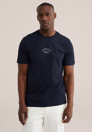 Mann trägt ein marineblaues kurzärmeliges T-Shirt mit weißem Schriftzug „Sunset State Canggu Afternoon“ und weiße Hose, steht vor neutralem Hintergrund.