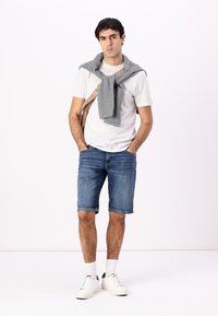 T-shirt blanche à col rond, pull gris noué au cou, shorts en denim, baskets blanches avec semelle à motifs et chaussettes blanches mi-mollet. Tenue minimaliste et décontractée.