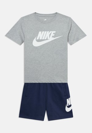 Svetlo siva kratka majica Nike z belim logotipom, kombinirana z temno modrimi kratkimi hlačami Nike z belim logotipom na levi nogi.