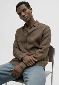 Brun linne långärmad skjorta med knapp-ner krage och ljusa knappar, tillsammans med blå denimjeans, sittande på en metallstol.