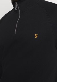 Sudadera negra con cuello de media cremallera, que presenta un pequeño logotipo amarillo bordado en el pecho. Fabricada en una suave tela texturizada.