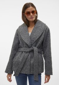 Manteau gris texturé avec un large col, taille cintrée et deux poches avant. Modèle portant des lunettes de soleil ambrées et un top blanc en dessous.