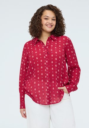 Kvinde med krøllet hår iført rød skjorte med lange ærmer og små paisley-mønstre og hvide bukser, smilende med den ene hånd i lommen.