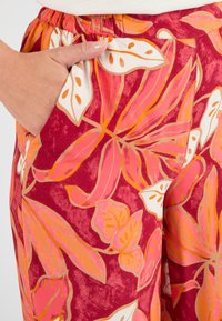 Pantaloni a fiori in rosso, rosa e panna, con vita elastica e tasche laterali. Presentano grandi foglie e dettagli accentuati.