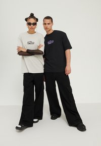Pair of models wearing oversized T-shirts; one in light gray with a logo and the other in black. Both wear black wide-leg pants and sunglasses.