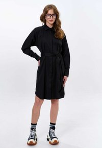 Schwarzes Hemdblusenkleid mit Kragen, langen Ärmeln und tailliertem Bund. Hergestellt aus glattem Stoff, verfügt es über eine durchgehende Knopfleiste und eine lockere Silhouette.