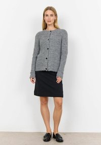 Grå stickad cardigan med texturerad tyg och svarta knappar, som bärs med en svart mönstrad kjol. Modellen har på sig svarta loafers.