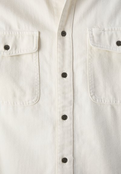 Camisa de mezclilla blanca con una tapeta de botones, dos bolsillos en el pecho y botones negros. La tela tiene un acabado texturizado.