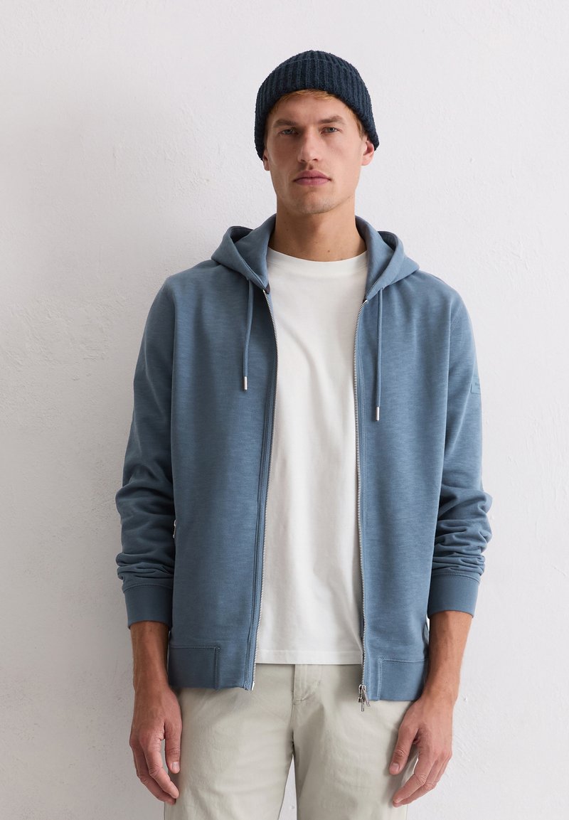 Hellblauer Kapuzenpullover mit Reißverschluss, glatter Textur, Kordelzugkapuze und gerippten Bündchen, kombiniert mit einem weißen T-Shirt und beigen Hosen.