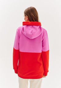 Mikina s barevnými bloky, která má růžovou horní část, červenou dolní část a překrývající se červenou kapuci. Vyrobena z hladké tkaniny s volným střihem.