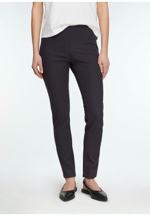 Kobieta ubrana w dopasowane czarne spodnie, biały top i czarne baleriny na jednolitym jasnym tle.