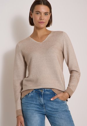 Jeune femme aux cheveux courts bruns, portant un pull beige clair à col en V et un jean bleu, debout avec une main dans la poche.