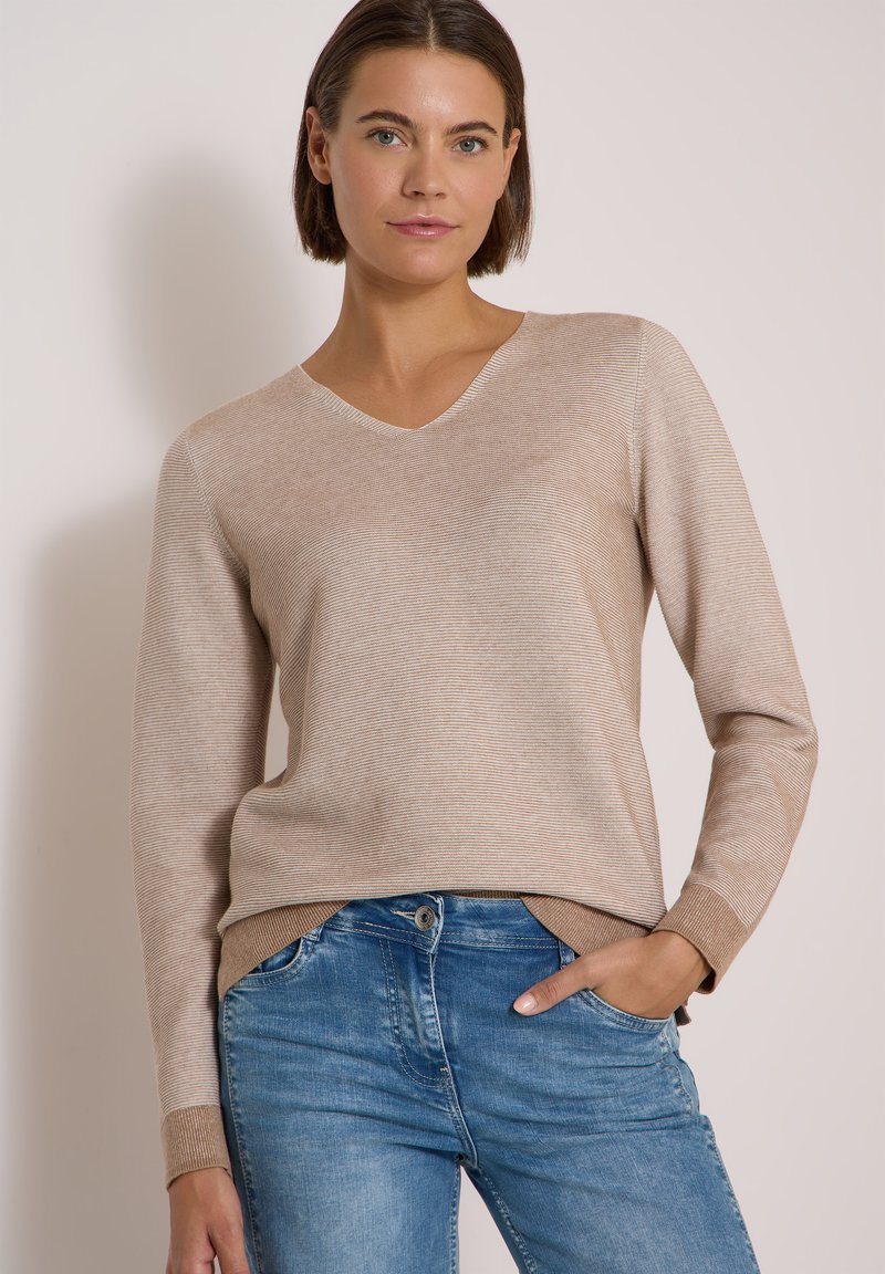 Jeune femme aux cheveux courts bruns, portant un pull beige clair à col en V et un jean bleu, debout avec une main dans la poche.