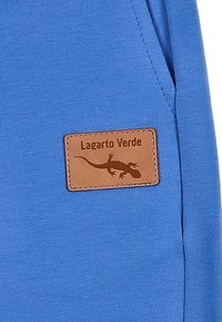 Blauwe stof met een gladde textuur, voorzien van een bruine leren patch met het label "Lagarto Verde" en een hagedis-silhouet.