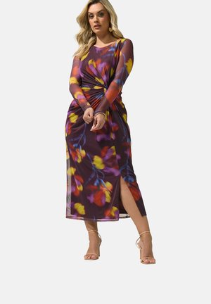 Woman wearing a long-sleeve, mid-length dress with a twisted front, floral pattern in purple, yellow, red, and blue, and a side slit.