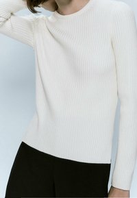 Prążkowana, długosłwowa, kremowa bluzka z miękkiego materiału, z okrągłym dekoltem; zestawiona z czarnymi spodniami, które mają dopasowany krój i fakturę.