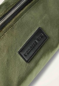 Pochette en tissu vert olive avec une fermeture éclair noire, ornée d'une étiquette rectangulaire en cuir noir embossée du nom "Vanessa Wu". Texture lisse.