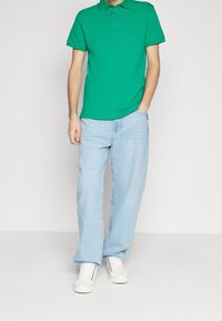 Polo vert en coton avec un col, associé à un jean en denim bleu clair et des baskets blanches. Comprend un petit logo sur la chemise.