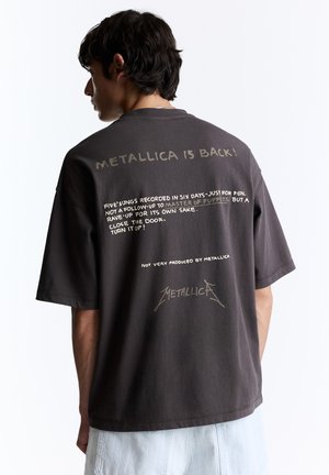 Jeune homme aux cheveux foncés portant un T-shirt noir Metallica avec un texte et un logo au dos, associé à un short de couleur claire.