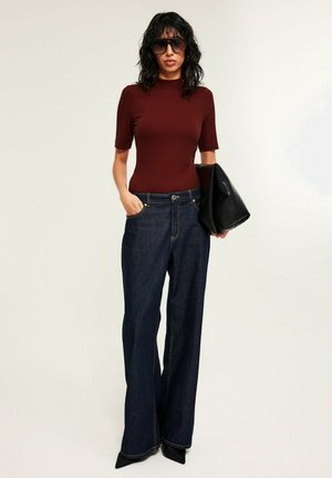 Kvinde iført mørkerød ribbet kortærmet top, mørkeblå jeans med vide ben, sorte spidse sko, stor sort clutch og store solbriller.