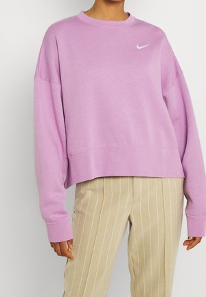 Sweatshirt Nike violet clair avec un col ras du cou, des épaules tombantes et des poignets côtelés. Associé à un pantalon beige à rayures.