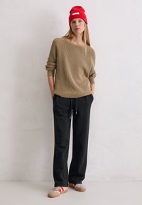 Vrouw in een beige gebreide trui, zwarte broek met trekkoord, beige en rode sneakers en een rode beanie, staand tegen een plain witte muur.