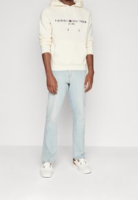 Kerman huppari, jossa on "TOMMY HILFIGER" -logo, yhdistettynä vaale blue-farkkuihin ja valkoisiin tennareihin, joissa on ruskeita yksityiskohtia. Yksinkertainen, rento asu.