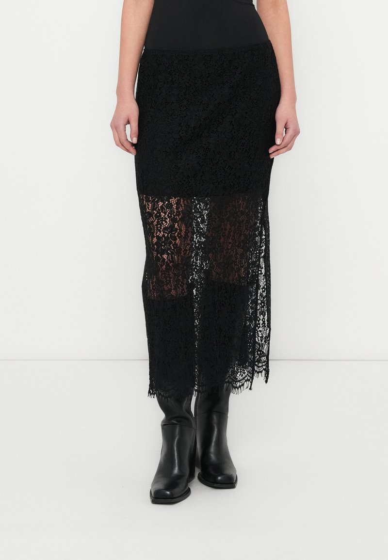 Sort midi-nederdel i blonder med gennemsigtigt design og en solid linning, fremhævet af en bølget kant, kombineret med knæhøje sorte støvler.