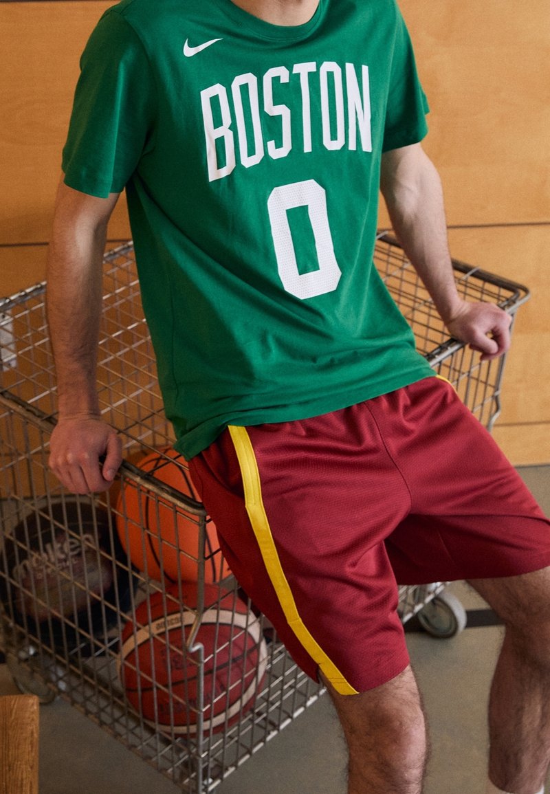 Jongeman in een groen Boston jersey en rode sportschoenen zit op een metalen kar en houdt basketballen vast binnenshuis.