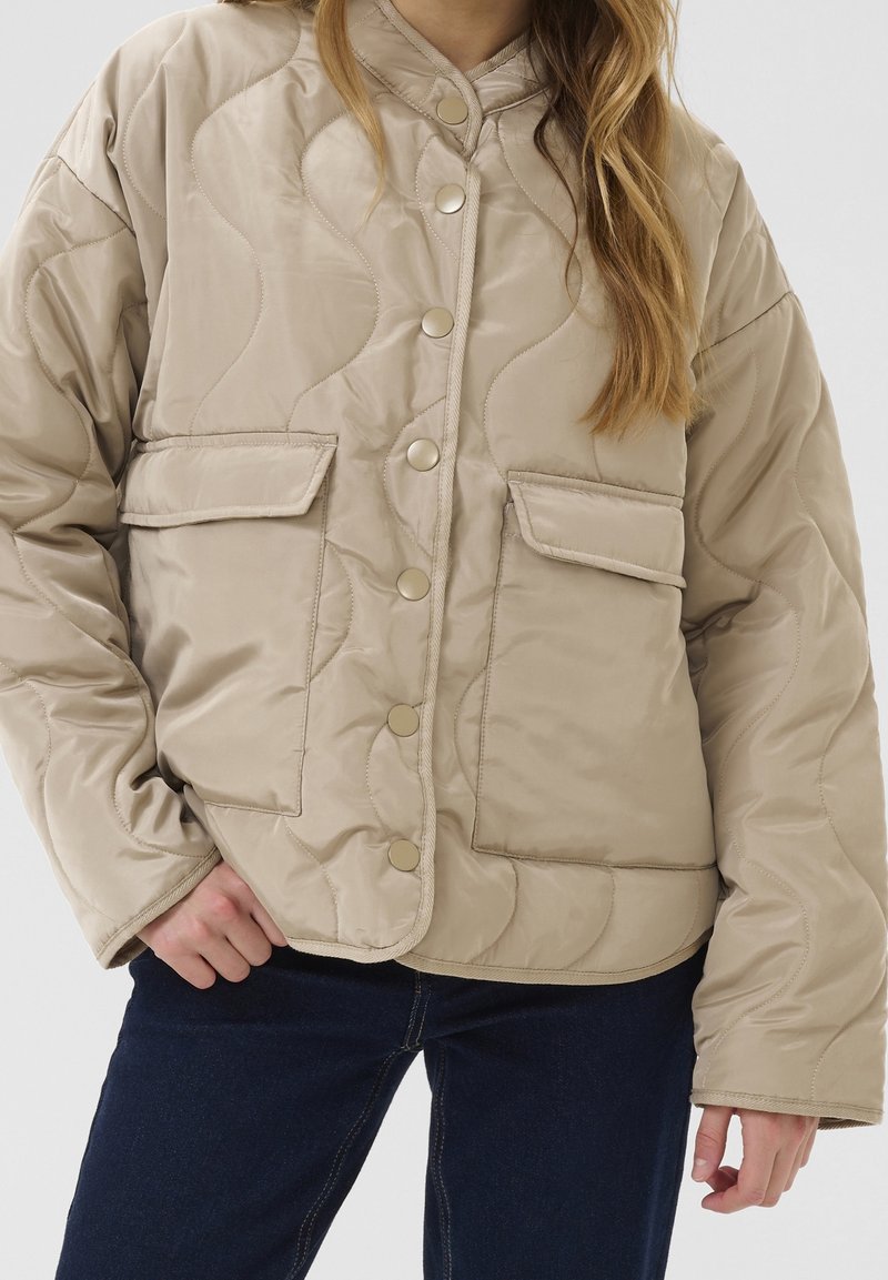 Veste matelassée beige avec boutons-pression et poches à rabat portée sur un jean bleu foncé par une personne aux longs cheveux châtain clair.