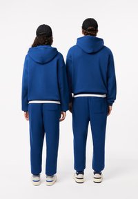Blauwe uniseks hoodie-sweatshirts hebben een relaxte pasvorm met een kangoeroezak. In combinatie met bijpassende blauwe sweatpants, die een zakdetail laten zien.