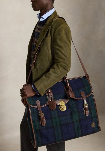 Uomo con blazer verde e maglione a fantasia porta una grande borsa scozzese blu e verde con tracolle in pelle e chiusura dorata.