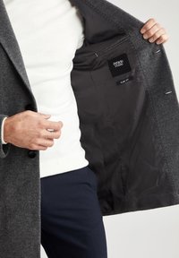 Grå uldblandingsfrakke med en notch-krave, to forreste knapper og en glat sort foring, der viser et slim fit-label.