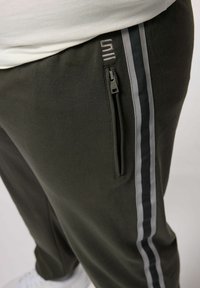 Donkergroene joggers met een ritsvak, voorzien van grijze strepen langs de zijkanten en een zachte, gestructureerde stof.